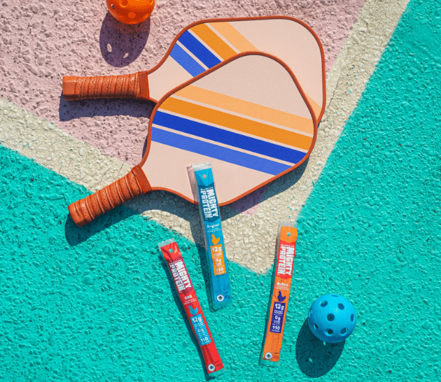 Des produits Mighty Protein sur un terrain de pickleball avec des raquettes.