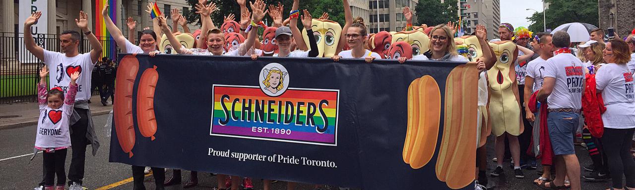 Employees marching in Pride Toronto parade holding Schneiders banner displaying pride colours.
