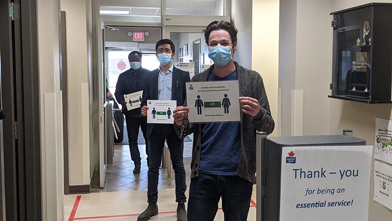 Trois employés portant des masques, tenant des panneaux sur la distanciation sociale à l'entrée de l'usine