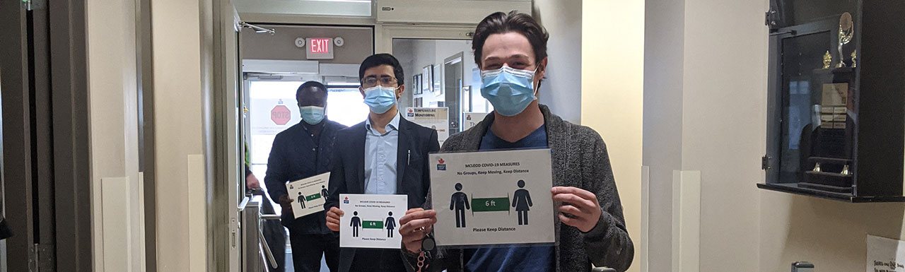 Trois employés portant des masques, tenant des panneaux sur la distanciation sociale à l'entrée de l'usine